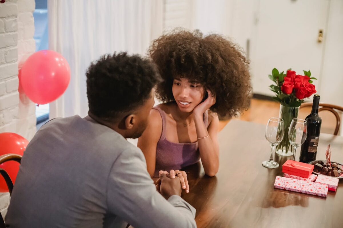 Young black couple in elegant clothes talking while holding hands at table with wine bottle and glasses near flowers bouquet in vase at light home while looking at each other